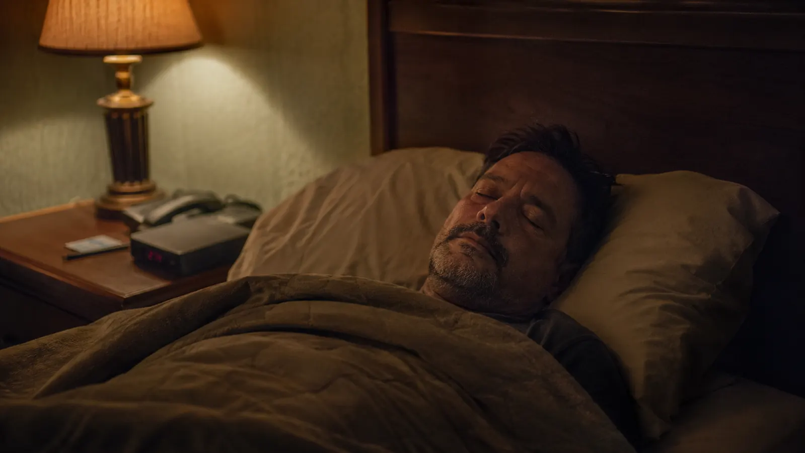 Man lying in a dark motel room under a heavy blanket, eyes closed, soft amber bedside lamp on, peaceful expression