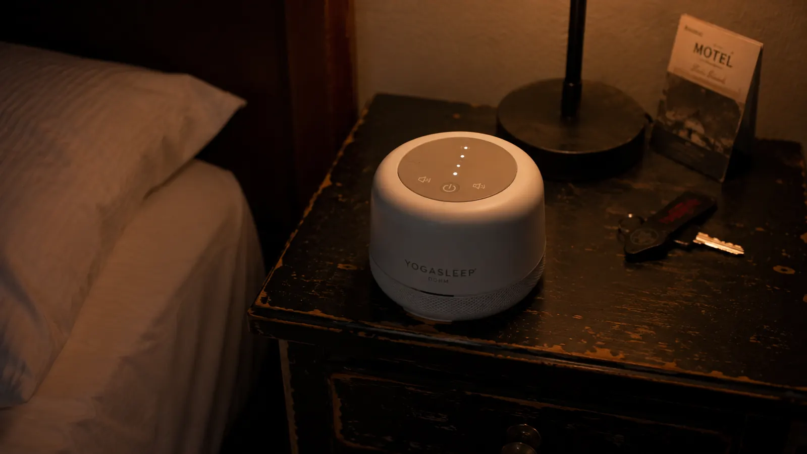 Yogasleep Dohm Classic white noise machine on a motel nightstand next to a glass of water, dim lamp light, night atmosphere